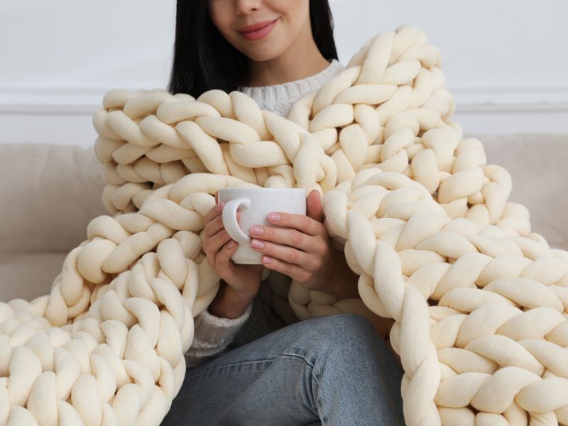 DIY blanket projects: a cozy chunky knit blanket in cream, wrapped around a woman enjoying a warm drink on a sofa